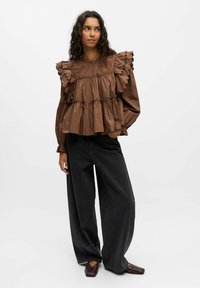 Blouse marron avec des détails froncés et festonnés, manches longues, et un design froncé, associée à un pantalon large noir et des chaussures marron.
