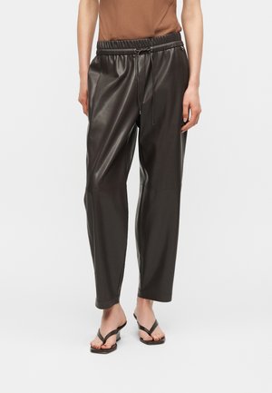 Woman wearing black faux leather drawstring pants with cropped legs and black heeled sandals, standing on white background.
