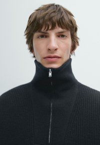 Svart stickad tröja med hög krage och halvdragkedja. Har en strukturerad tygkvalitet och subtila ribbade detaljer längs sömmarna.