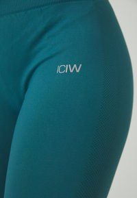 Tirkizne tajice s teksturiranim bočnim panelom i glatkom površinom, s malim, bijelim "ICIW" logotipom na lijevom bedru.