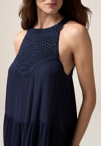 Rochie fără mâneci de culoare bleumarin, cu detalii din croșetă la decolteu, țesătură texturată și un tiv cu volane.