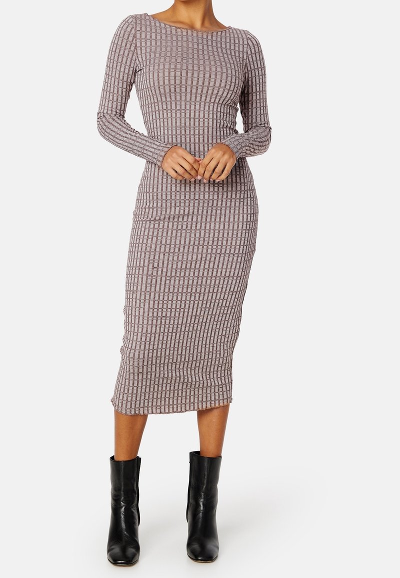 Femme portant une robe midi ajustée à manches longues avec un motif à carreaux et des bottines noires, debout devant un fond uni.