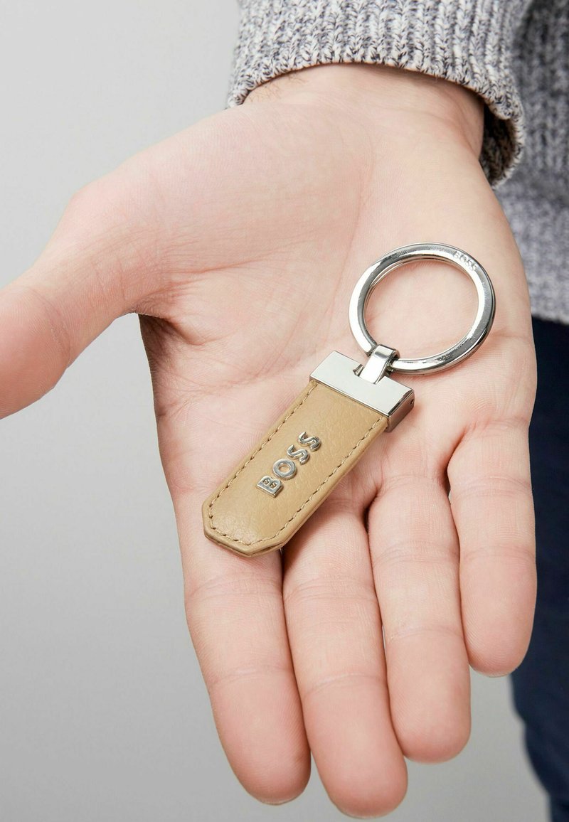 Portachiavi in pelle beige, con un anello in metallo argento e un pendente rettangolare con la scritta "BOSS" in rilievo. Texture piatta e liscia.
