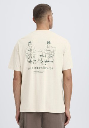 Mand iført lysebeige T-shirt med illustration af to mænd iført solbriller, der sidder på stole, og teksten "Out Of Office Since '99".