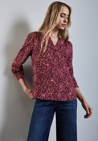 Blouse à motif bourgogne avec décolleté en V, manches longues et coupe décontractée. Le design présente des formes abstraites roses sur un tissu lisse.