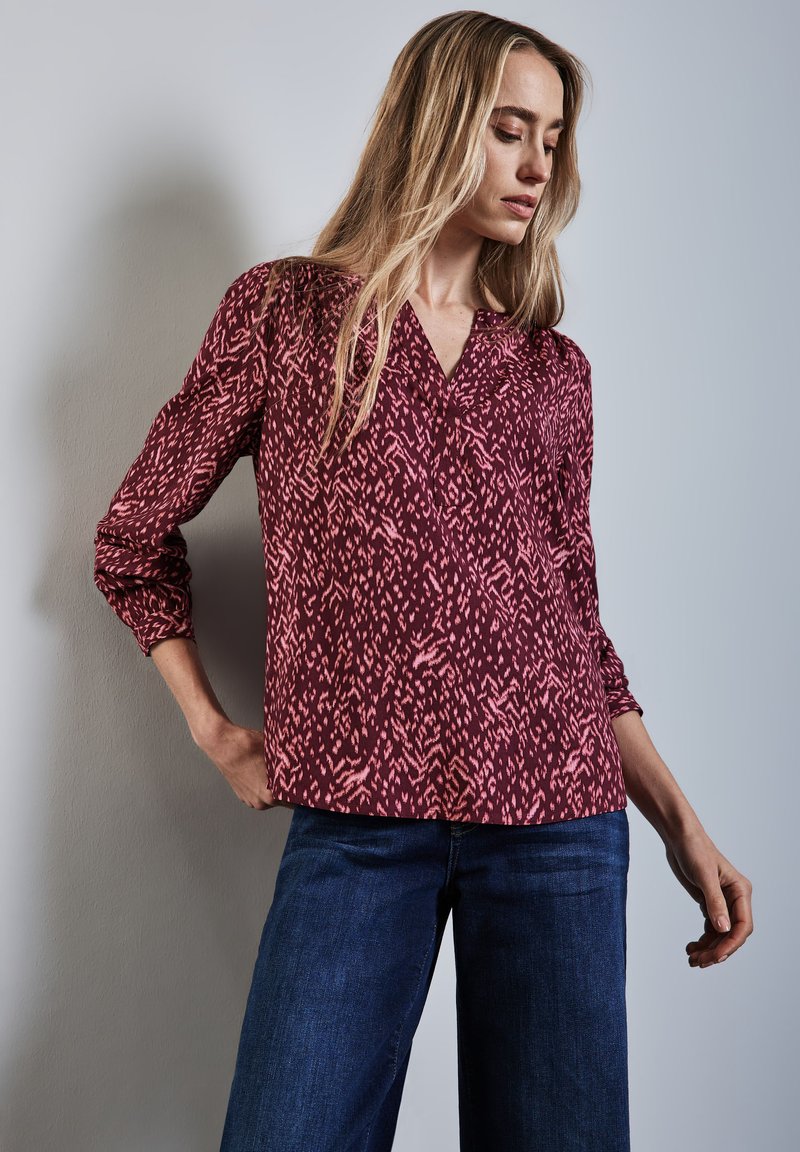 Blouse à motif bourgogne avec décolleté en V, manches longues et coupe décontractée. Le design présente des formes abstraites roses sur un tissu lisse.