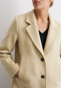 Beiger Wollmantel mit Reverskragen, zwei vorderen Taschen und zwei schwarzen Knöpfen. Weiche Textur mit auffälligen Nähdetails.