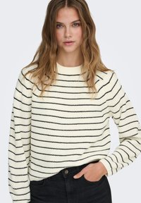 Pull crème avec des rayures horizontales noires, texture en tricot, et un col rond. Associé à un jean noir, mains dans les poches, coupe décontractée.