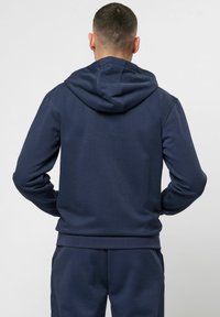 Tumesinane kapuutsiga ziipiga sviiter, mis on valmistatud pehmest kangast, millel on nööriga kapuuts, soonikkoes mansetid ja alläär. Sile tekstuur, minimalistlik disain, lõdva lõikega.