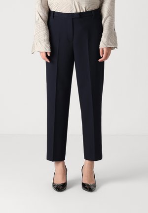 Donna che indossa pantaloni blu navy sartoriali a gamba dritta, top a maniche lunghe testurizzato color avorio e décolleté nere in vernice.