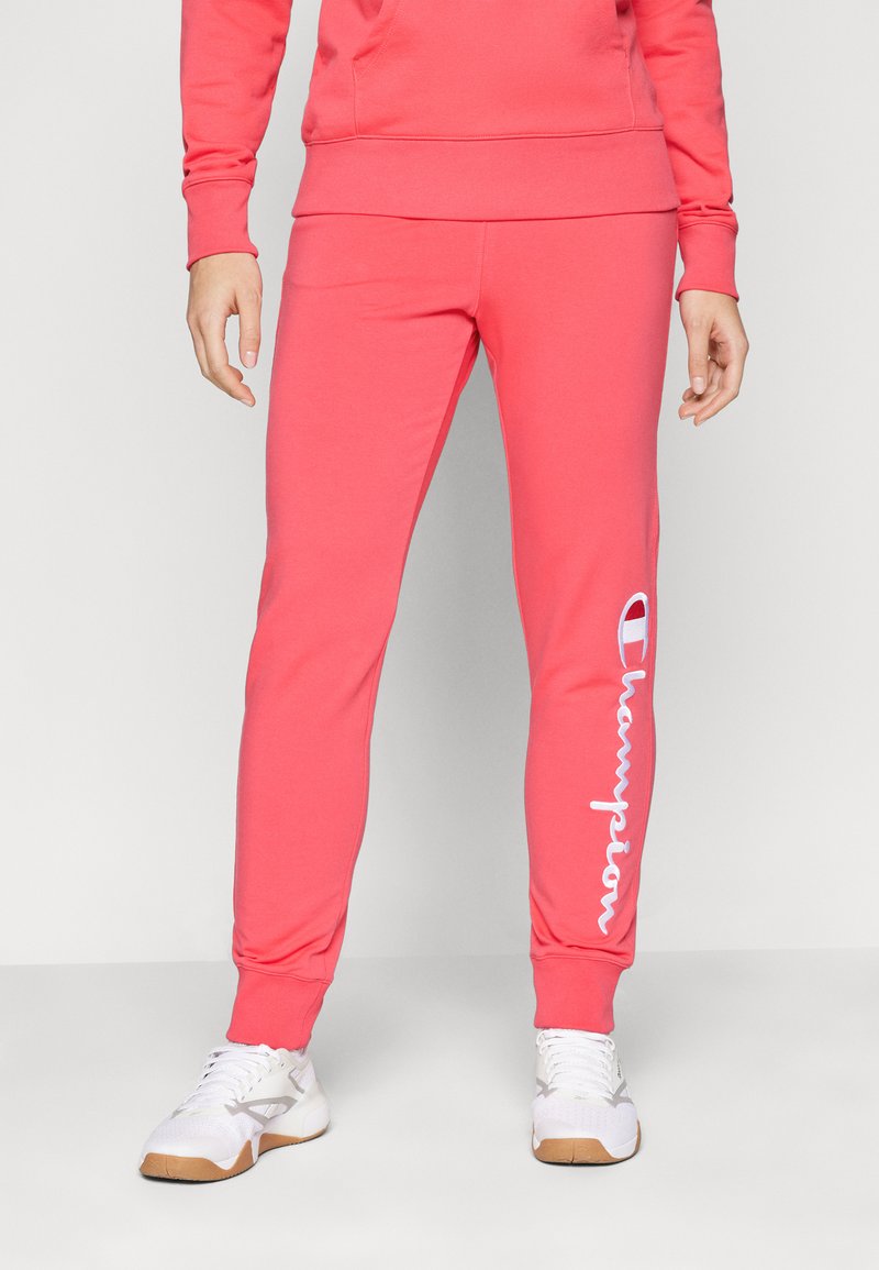 Pantaloni de trening roz, fabricați dintr-o țesătură moale, cu un design conic. Prezintă un logo alb "Champion" pe piciorul stâng și manșete elasticizate.