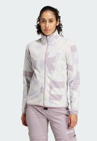 Chaqueta de forro polar en púrpura claro con patrones geométricos, cierre frontal de cremallera, cuello alto y detalle del logotipo. Combinada con pantalones a juego en púrpura claro.