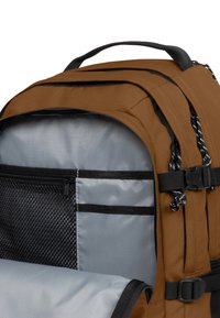 Mochila de tela marrón con interior gris, varios compartimentos, un bolsillo de malla con cremallera y correas ajustables con detalles en negro.
