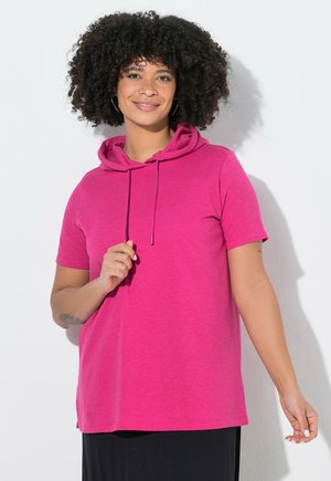 Kobieta z kręconymi włosami, ubrana w jasnoróżową bluzę z krótkim rękawem z kapturem, uśmiechnięta, stojąca przy białej ścianie z cieniami.