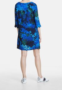 Květinové šaty s modrými, zelenými a námořnickými vzory; tříčtvrteční rukávy a volný střih. V kombinaci s bílými teniskami s dekorativními detaily.