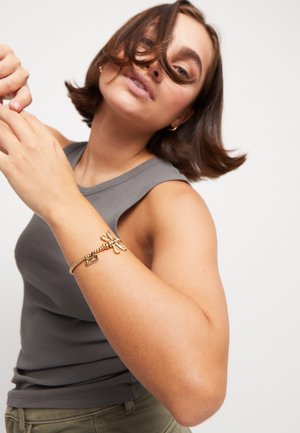 Pulsera de charms de oro con un diseño de libélula, que presenta una cadena texturizada y una etiqueta rectangular, llevada en un brazo desnudo con una camiseta de tirantes gris.