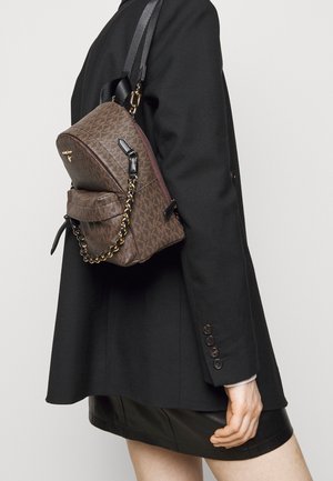 Zaino in nylon marrone con motivo, hardware dorato, elemento a catena e spallacci regolabili. Indossato sulla schiena, abbinato a un blazer nero.