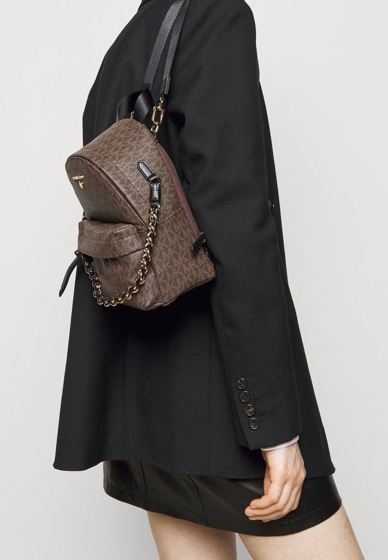 Zaino in nylon marrone con motivo, hardware dorato, elemento a catena e spallacci regolabili. Indossato sulla schiena, abbinato a un blazer nero.