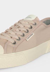 Baskets en toile beige Calvin Klein avec semelle en caoutchouc crème, œillets métalliques, lacets beiges et petite étiquette logo Calvin Klein sur le côté.
