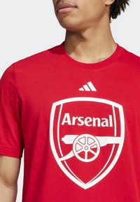 Červené bavlnené tričko s bielym logom Arsenal a emblémom dela, okrúhly výstrih a krátke rukávy. Logo značky na pleci.