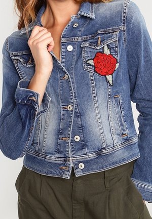 Femme portant une veste en jean bleu délavé avec une broderie de rose rouge sur la poche poitrine et un pantalon vert olive, main tenant le col de la veste.
