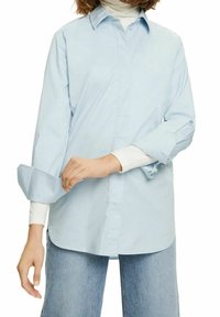 Ljusblå skjorta med knappar, långa ärmar och uppvikta manschetter, tillverkad av mjukt material, i kombination med en vit polotröja och blå jeans.