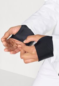 White long-sleeve garment with black satin cuffs; hands adjusting cuffs. Smooth texture, tailored fit, and contrasting color details.