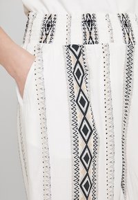 Ruka v kapse bílých kalhot s černými a béžovými vertikálními geometrickými a cik-cak vzory, elastický pas.