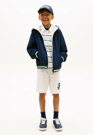 Smilende dreng iført navyblå kasket, navyblå lynlåstrøje, hvide polo med striber, hvide shorts med grønt logo, navyblå sokker og navyblå sneakers.