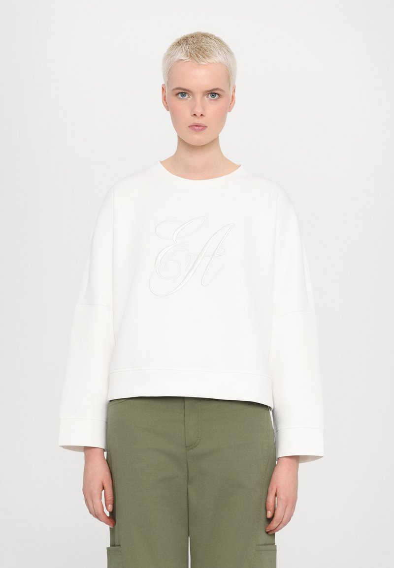 White sweatshirt with a cropped design, featuring drop shoulders and an embroidered logo. Worn with olive green cargo pants.