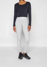 Schwarzes, kurzes Langarmoberteil mit weißen Nähten, kombiniert mit hellgrauen, hoch taillierten Leggings und schwarzen Sportschuhen.