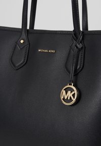 Sac fourre-tout en cuir noir à finition texturée, avec ferrures dorées, logo embossé et double poignées supérieures. Détail de couture distinctif sur les poignées.