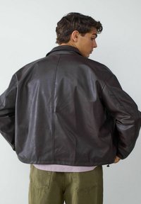 Bruine leren jas met een ontspannen pasvorm, brede kraag en zichtbare naaddetails, gecombineerd met een lichtroze top en olijfgroene broek.