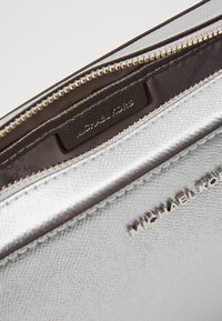 Monedero de textura plateada de Michael Kors con cremallera en tono dorado parcialmente abierta, mostrando el tejido interior con la marca y la etiqueta de cuero.