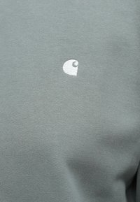 Szürke pamut póló, bal oldalon egy kis fehér logóval. Az anyag sima textúrájú, egyszínű kivitelű.