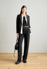 Černý blazer přes bílou košili a černý top, doplněný širokými černými kalhotami. Doplňky zahrnují úzký černý pásek a malou černou tašku.