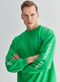 Zielony bawełniany sweter z długimi rękawami. Posiada niebieski napis "PARK" oraz "CASUAL CLUB" na lewym rękawie. Klasyczny krój z okrągłym dekoltem.
