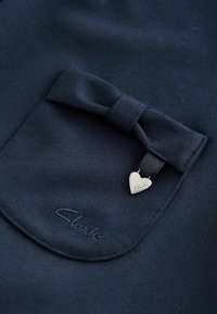 Marinfärgad tyg med texturerad yta har en liten ficka med en rosett och en hjärtformad silverberlock med texten "Clark" präglad.