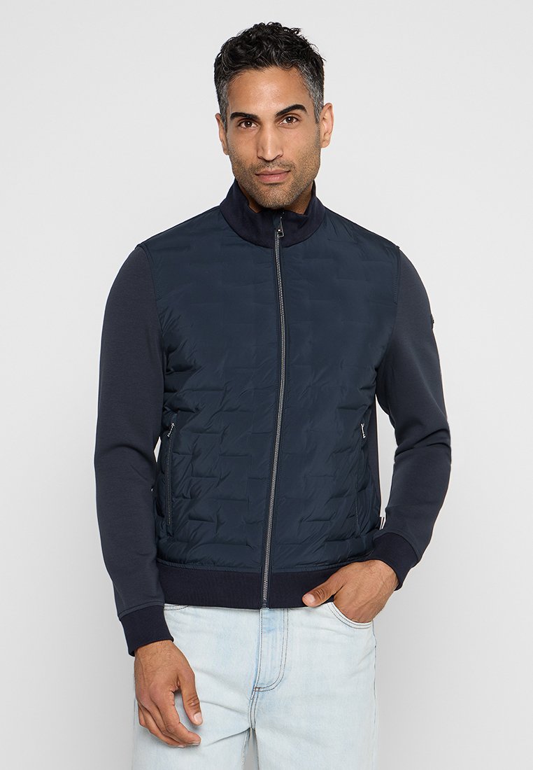 Gilet trapuntato navy con colletto alto e chiusura zip, dotato di maniche in maglia e orlo a costine. Texture liscia con dettagli di motivo sottili.