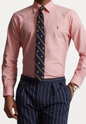 Mężczyzna noszący różową koszulę na guziki, granatowe spodnie w paski, granatowy krawat z żółtymi ukośnymi paskami i małymi herbami oraz zegarek na rękę.