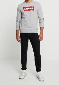 Sudadera gris con logo rojo de "Levi's", puños y dobladillo de canalé, combinada con jeans negros ajustados y zapatillas blancas.