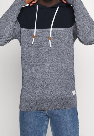 Man die een marineblauwe en grijze gebreide hoodie met witte trekkoorden verstelt, draagt blauwe jeans, tegen een effen lichte achtergrond.