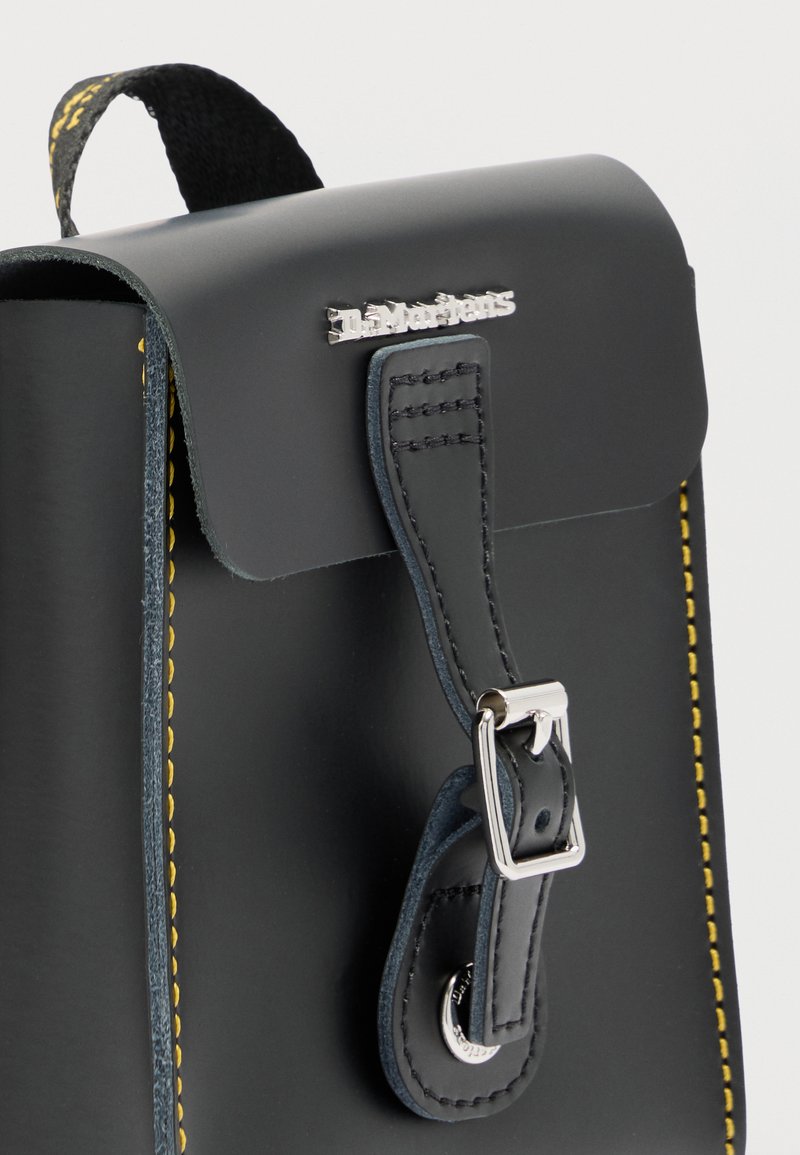Borsa Dr. Martens in pelle nera con cinturino frontale con fibbia e cuciture gialle lungo i bordi su sfondo bianco.