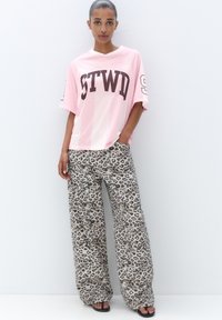 Růžová síťovaná oversized košile s nápisem "STWD" v tmavě hnědé. Volné leopardí kalhoty s širokými nohavicemi. Černé sandály. Jednoduché bílé pozadí.