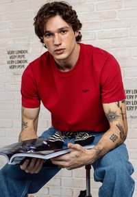 Rdeča bombažna majica s ovratnikom, črn logotip na prsih. Modri jeans. Model sedi in drži revijo, z vidnimi tetovažami.
