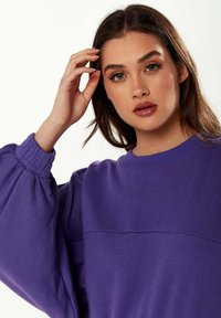 Purple sweatshirt with a textured fabric, rounded neckline, and puffed sleeves. Features a seam detail across the chest.
