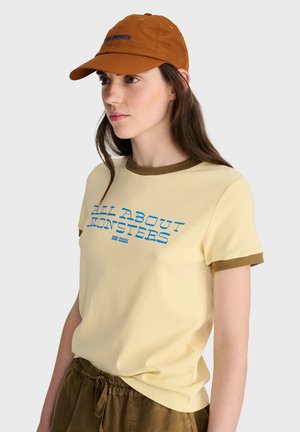 Jeune femme portant une casquette orange, un t-shirt jaune avec un texte bleu "All About Monsters," et un short marron, regardant sur le côté sur un fond clair.