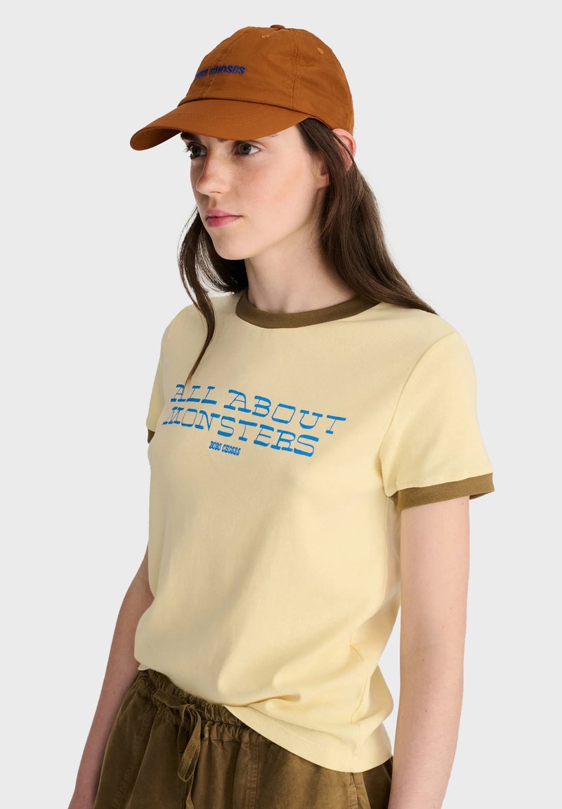 Jeune femme portant une casquette orange, un t-shirt jaune avec un texte bleu "All About Monsters," et un short marron, regardant sur le côté sur un fond clair.