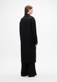 Abrigo largo negro con un diseño de cuello, hecho de una tela texturizada. Corte holgado, llega a la mitad de la pantorrilla, llevado sobre pantalones amplios.