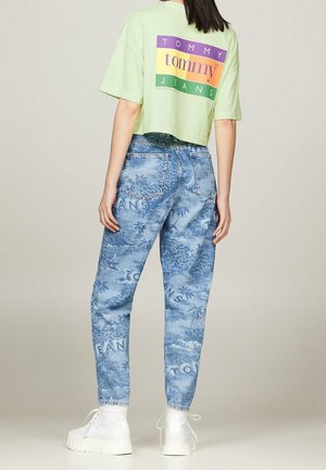 Vrouw draagt een lichtgroen cropped T-shirt met Tommy Jeans-logo en blauwe patroon spijkerbroek met witte veterlaarzen, staand met de rug naar voren.